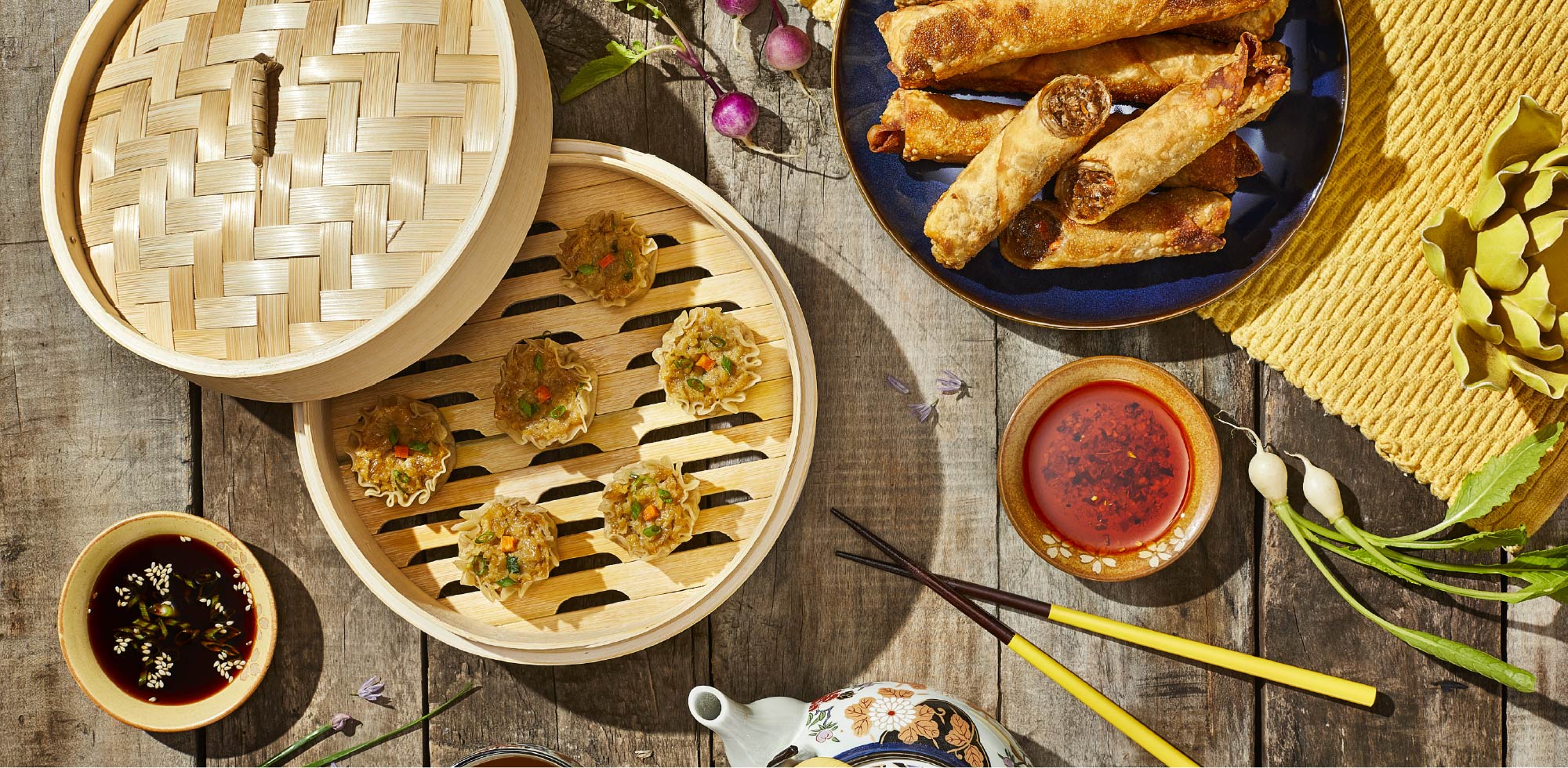 A table is set with a spread of egg rolls, steamed dumplings, dipping sauces, and chopsticks